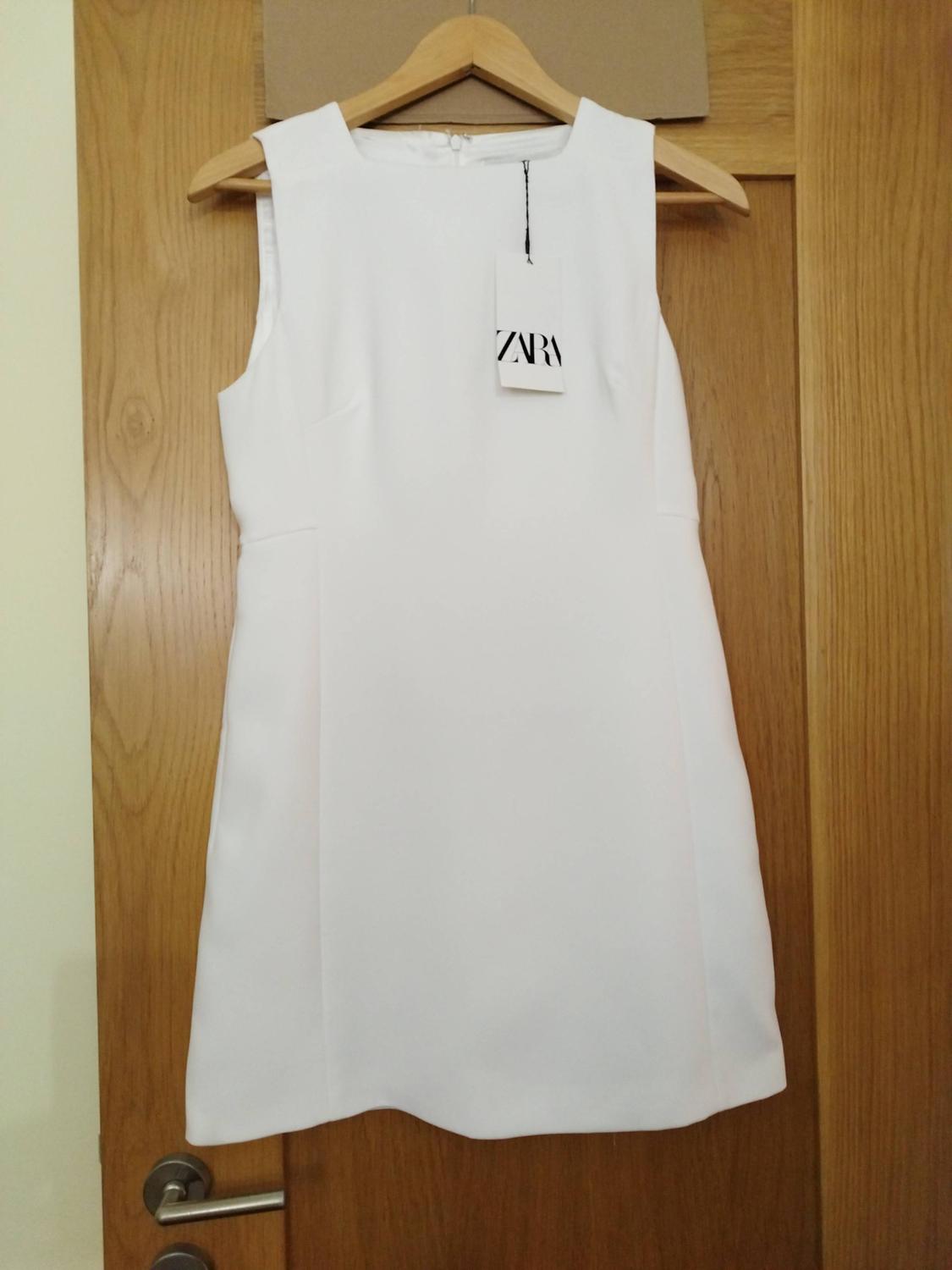 Short white sleeveless dress hanging on a hanger on a wooden door. Visible label with the text ZARA.