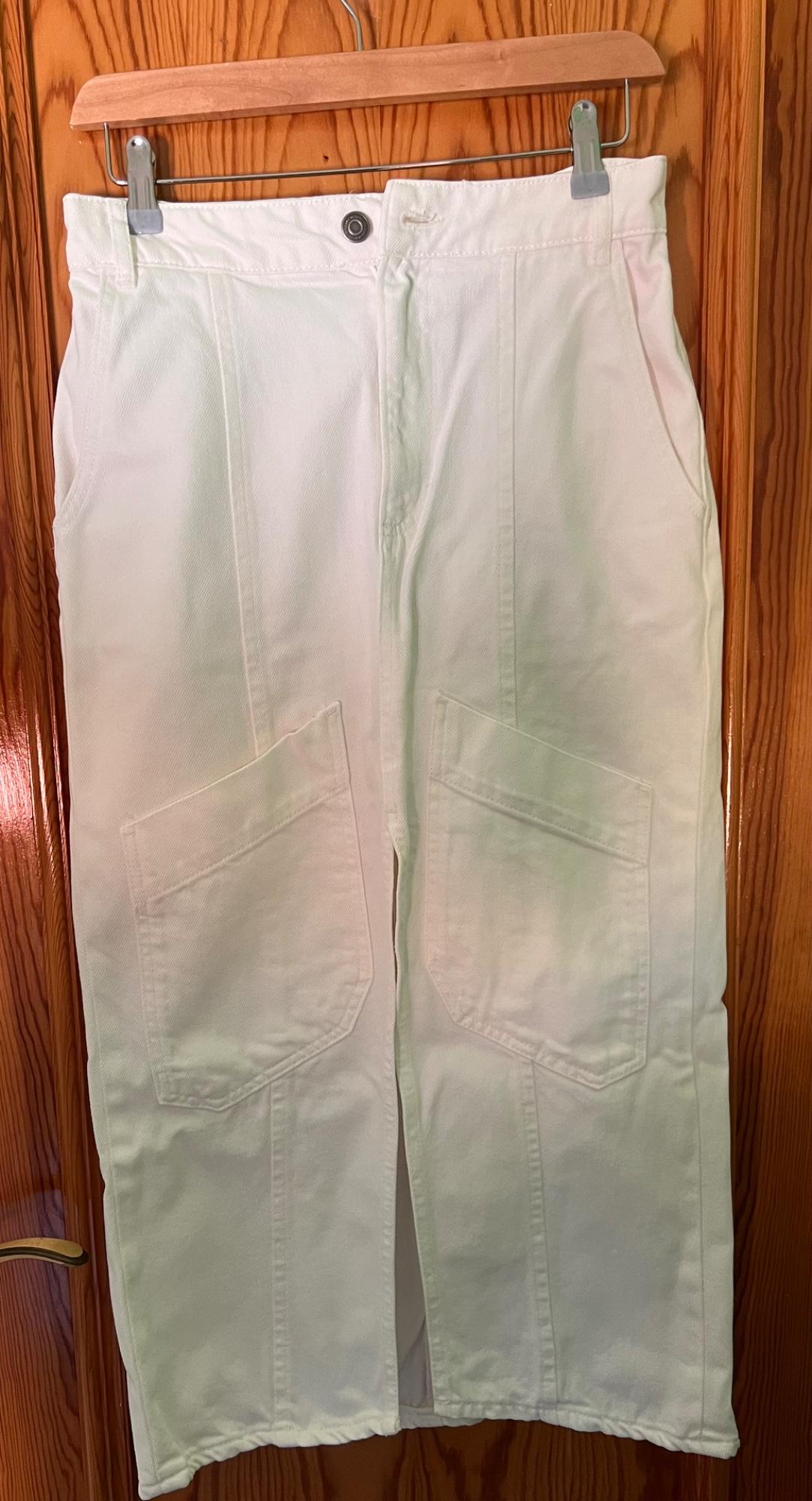 White trousers hanging on a wooden hanger, with large pockets at the front, against a wooden door background.