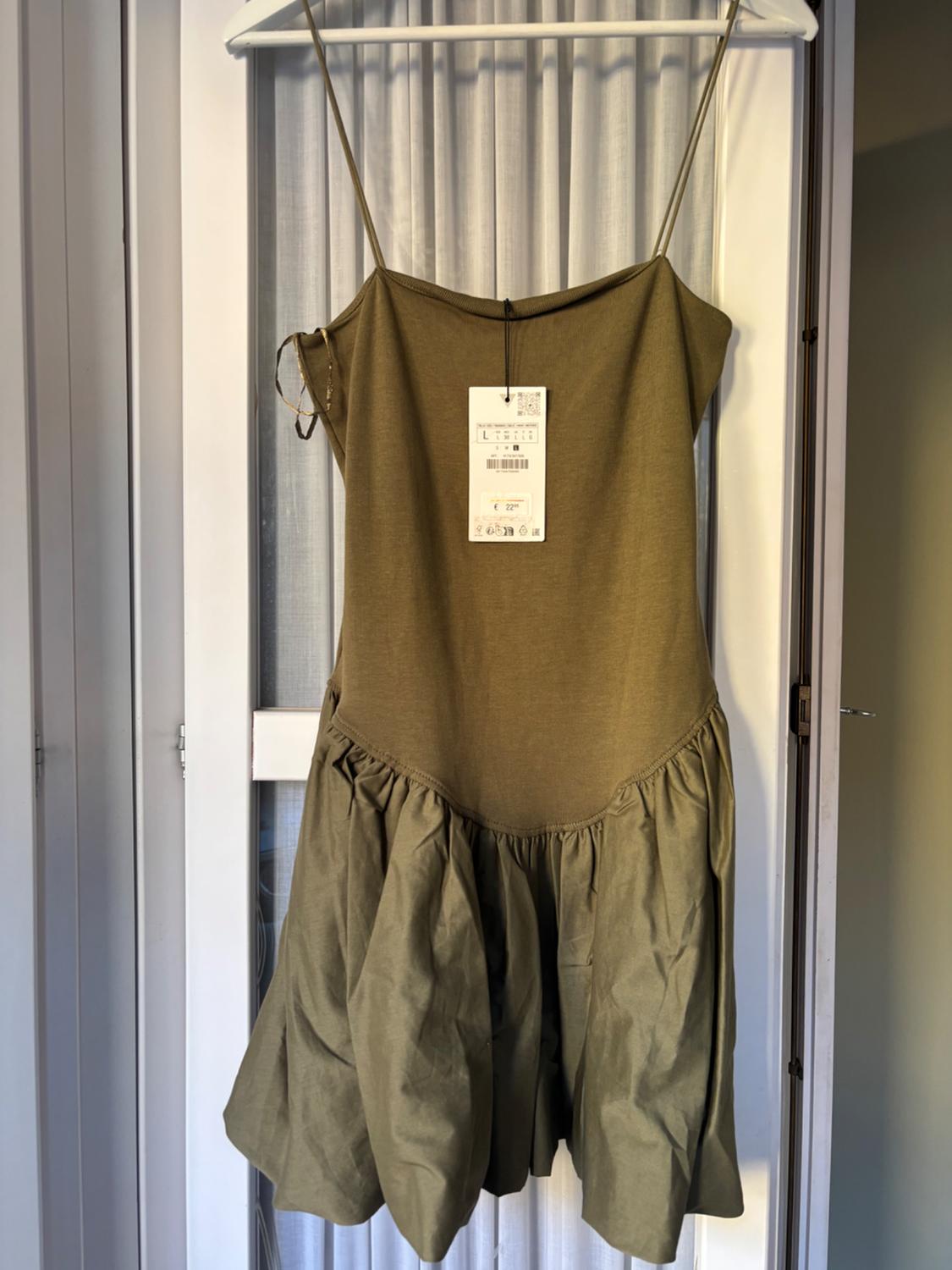 Olive green dress with spaghetti straps, visible hanging tag, hanging on a coat hanger in front of a white curtain and glass door.