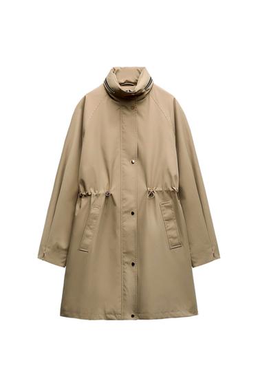 Beige coat with a high collar and adjustable drawstring waist. Front pockets and snap button closure. White background.