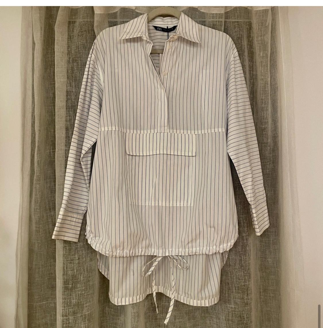 Black and white vertical striped shirt hanging on a hanger, with a front pocket and drawstring hem. Beige curtain background.