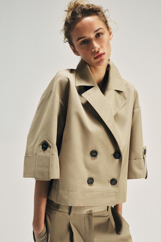 Střední detail opékaného trenčkotu s klopovým límcem, tříčtvrtečními rukávy s páskem a knoflíkem, dvouřadým zapínáním na knoflíky.