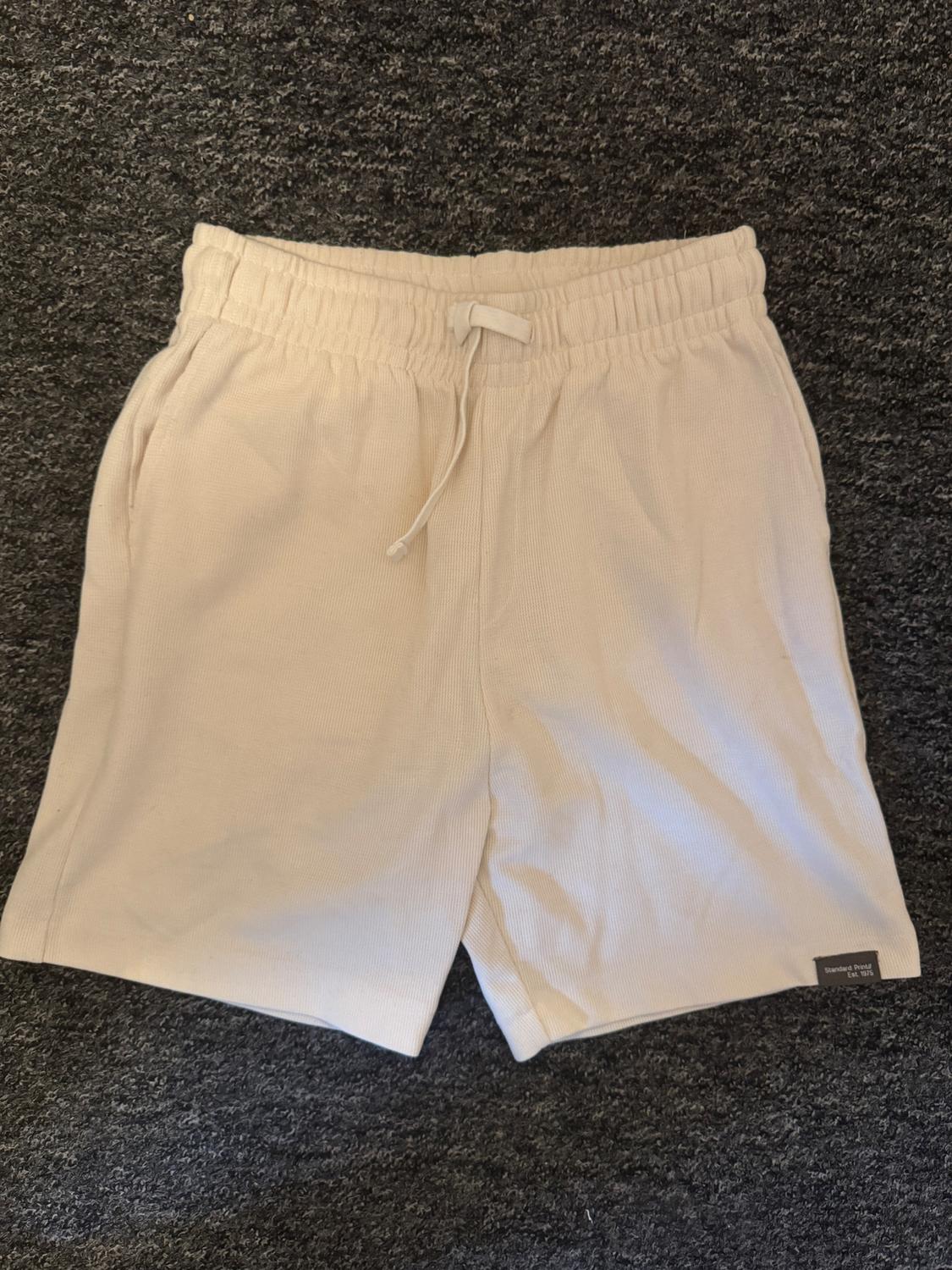 Beige shorts with an elasticated waist and adjustable drawstring, on a dark grey background. Visible label on the bottom right.