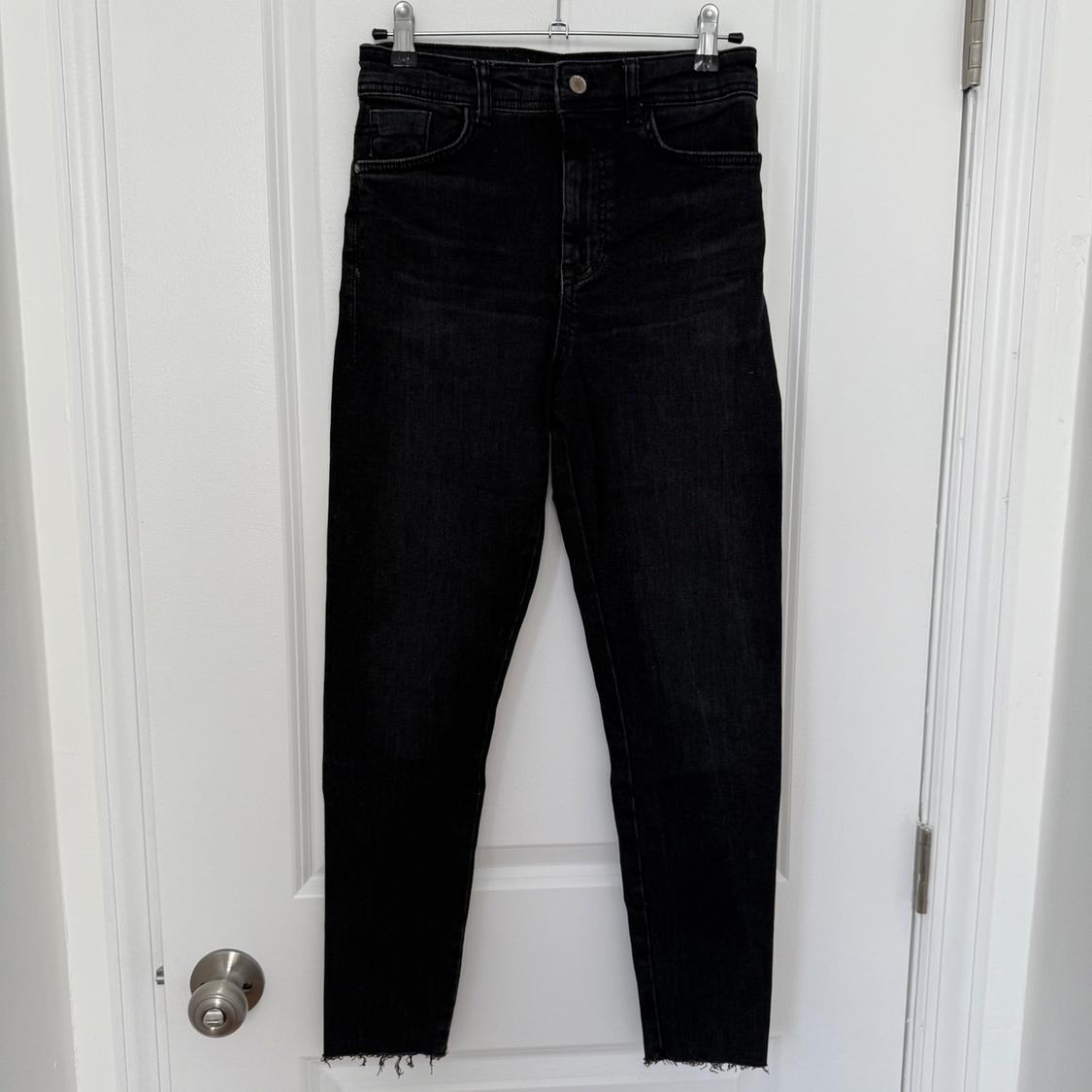 Black denim pants with a slim fit and frayed hem, hanging on a white door with a silver doorknob.