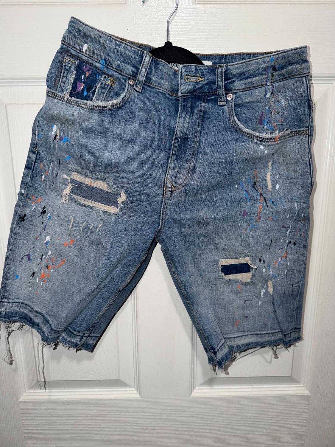 Blue denim shorts with a distressed effect and colourful paint splatters, hanging on a white door.