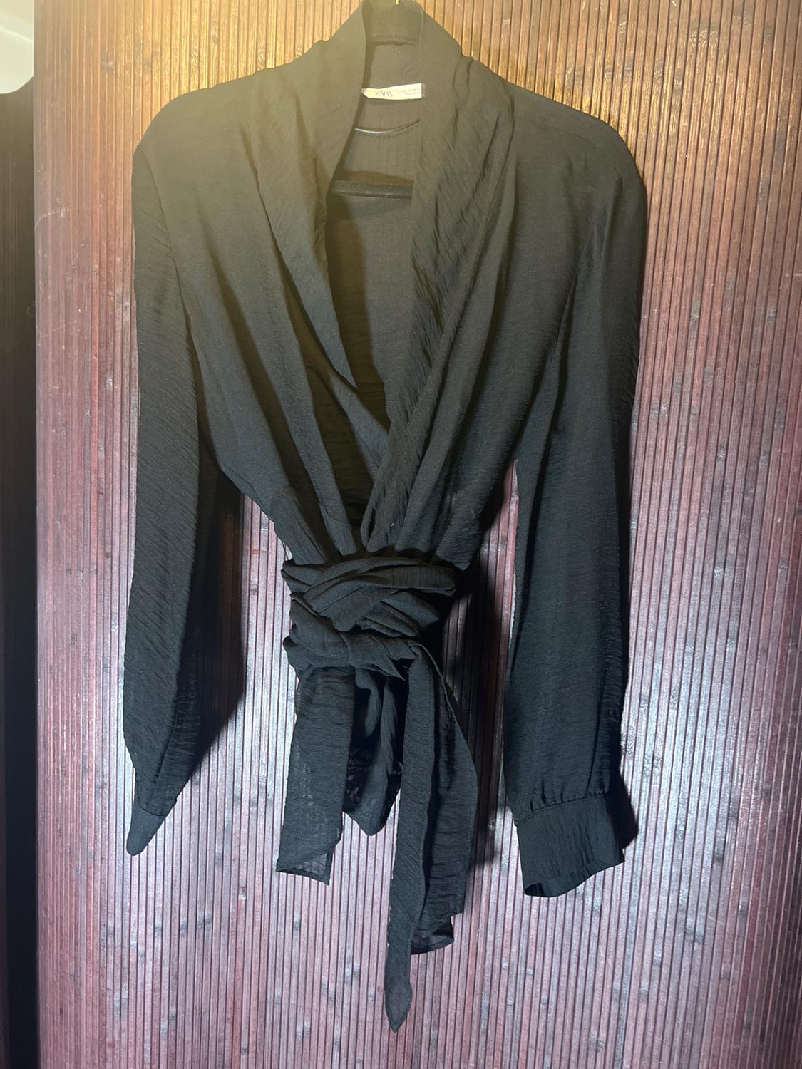Black long-sleeved blouse with a draped neckline and tie belt, hanging on a hanger against a brown wooden background.