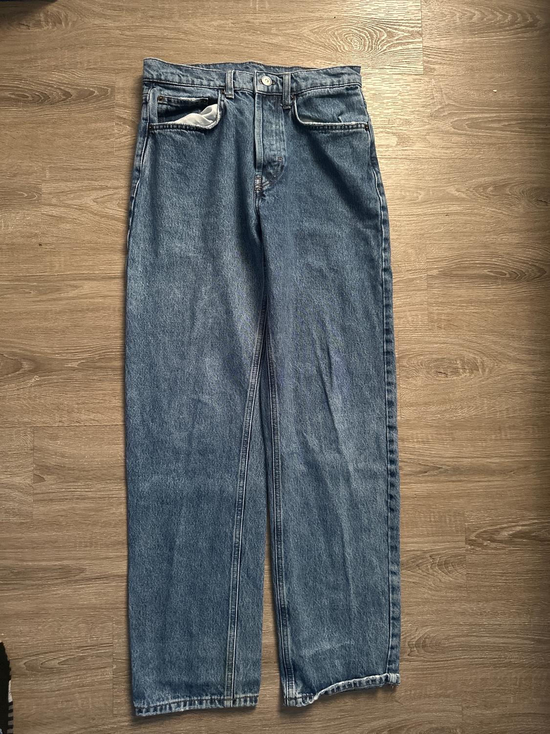 Straight-cut blue jeans on a light wooden floor. Visible front pockets, one partially turned inside out.