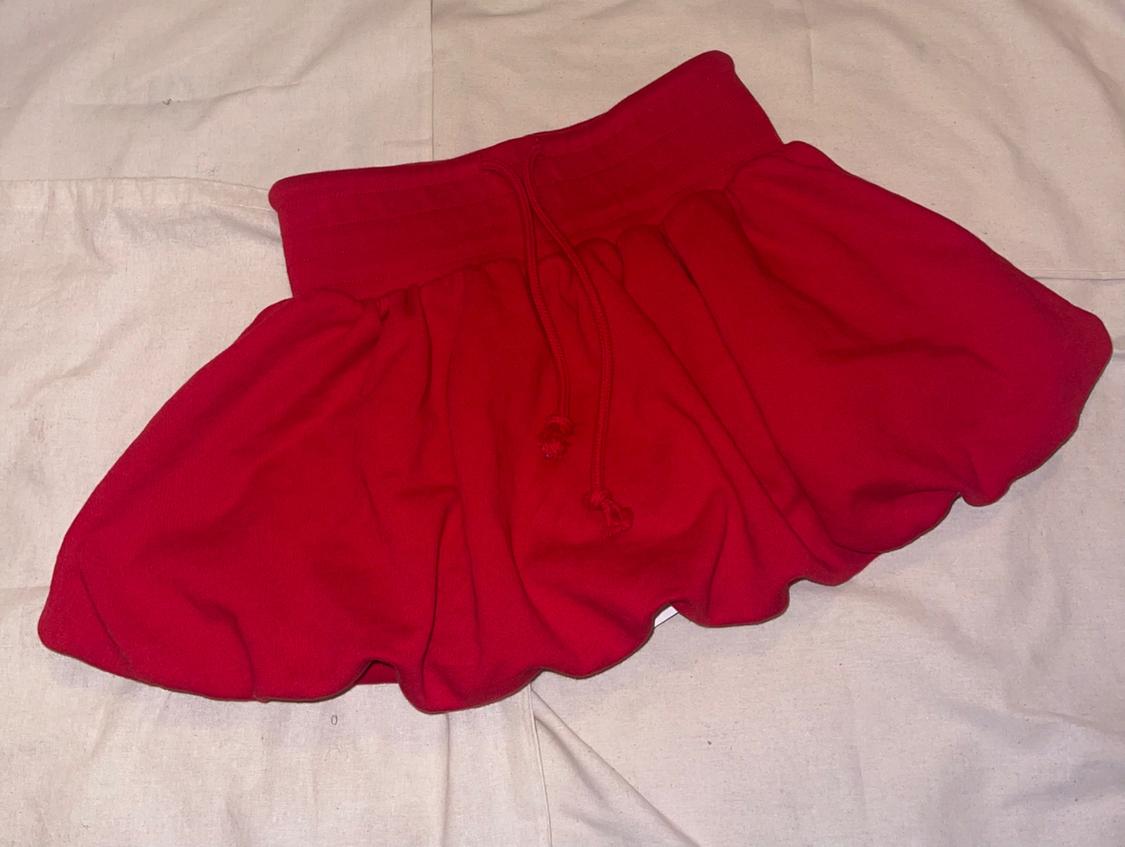 Red short balloon-style skirt with an elastic waistband and drawstring, placed on a light wrinkled fabric with a casual and relaxed style.