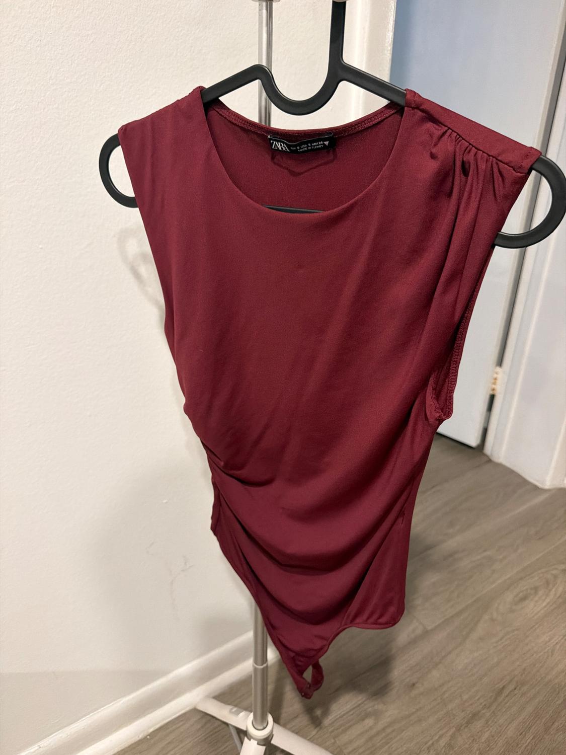 Garnet sleeveless ruched bodysuit, hanging on a black hanger on a metal stand. White wall, wooden floor. Text: ZARA.