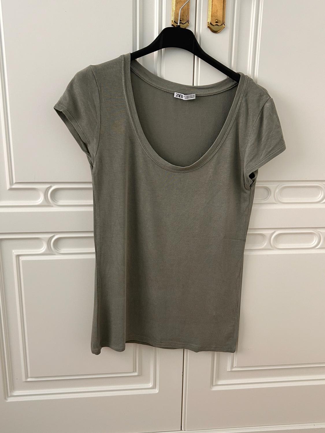 Green short-sleeved round-neck T-shirt hanging on a black hanger in front of a white door. Label with the text ZARA.