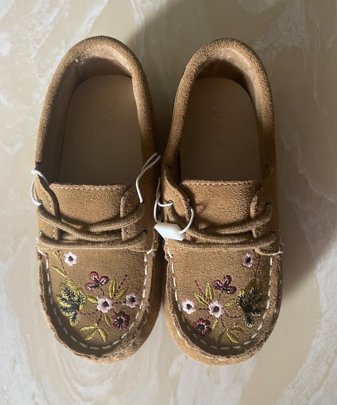Brown suede shoes with floral embroidery in shades of green, pink, and yellow, on a light background.