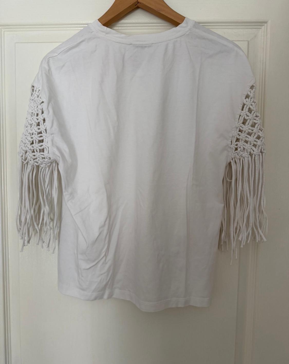 Λευκό T-shirt κρεμασμένο σε ξύλινη κρεμάστρα, με μανίκια από μακραμέ και μακριά κρόσσια, σε φόντο λευκής πόρτας.