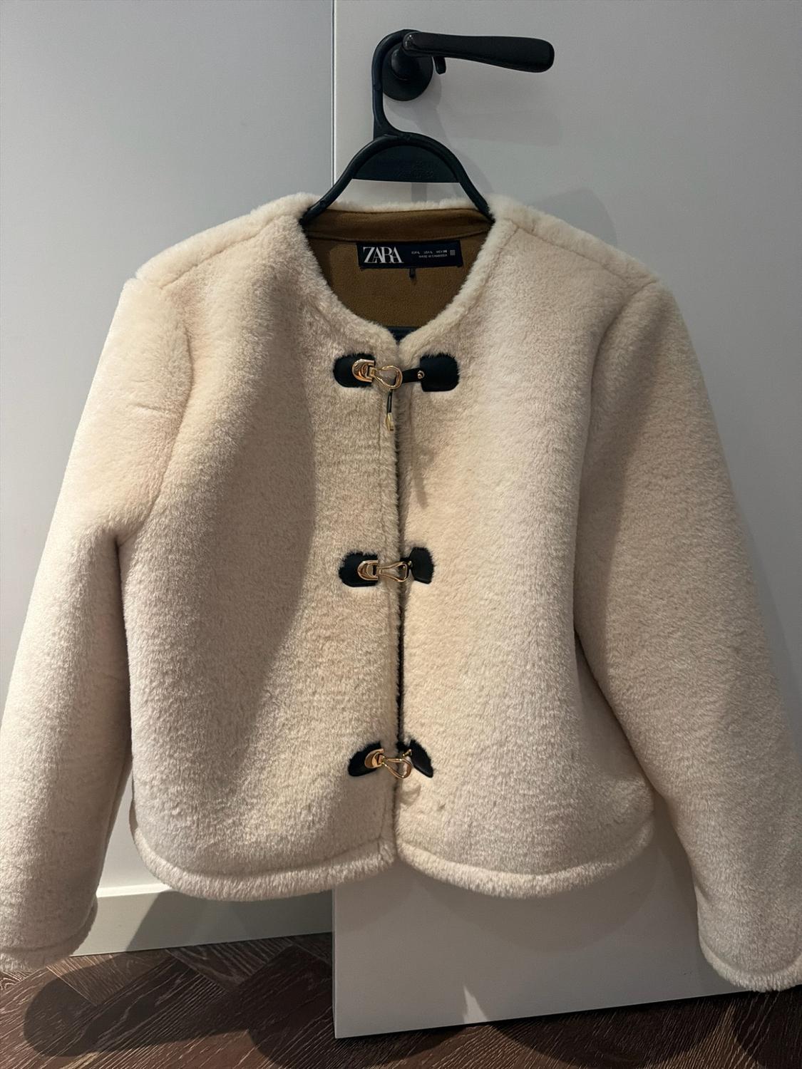 Soft-textured cream jacket hanging on a hanger. It has three black fastenings with gold details and the ZARA label on the inside.