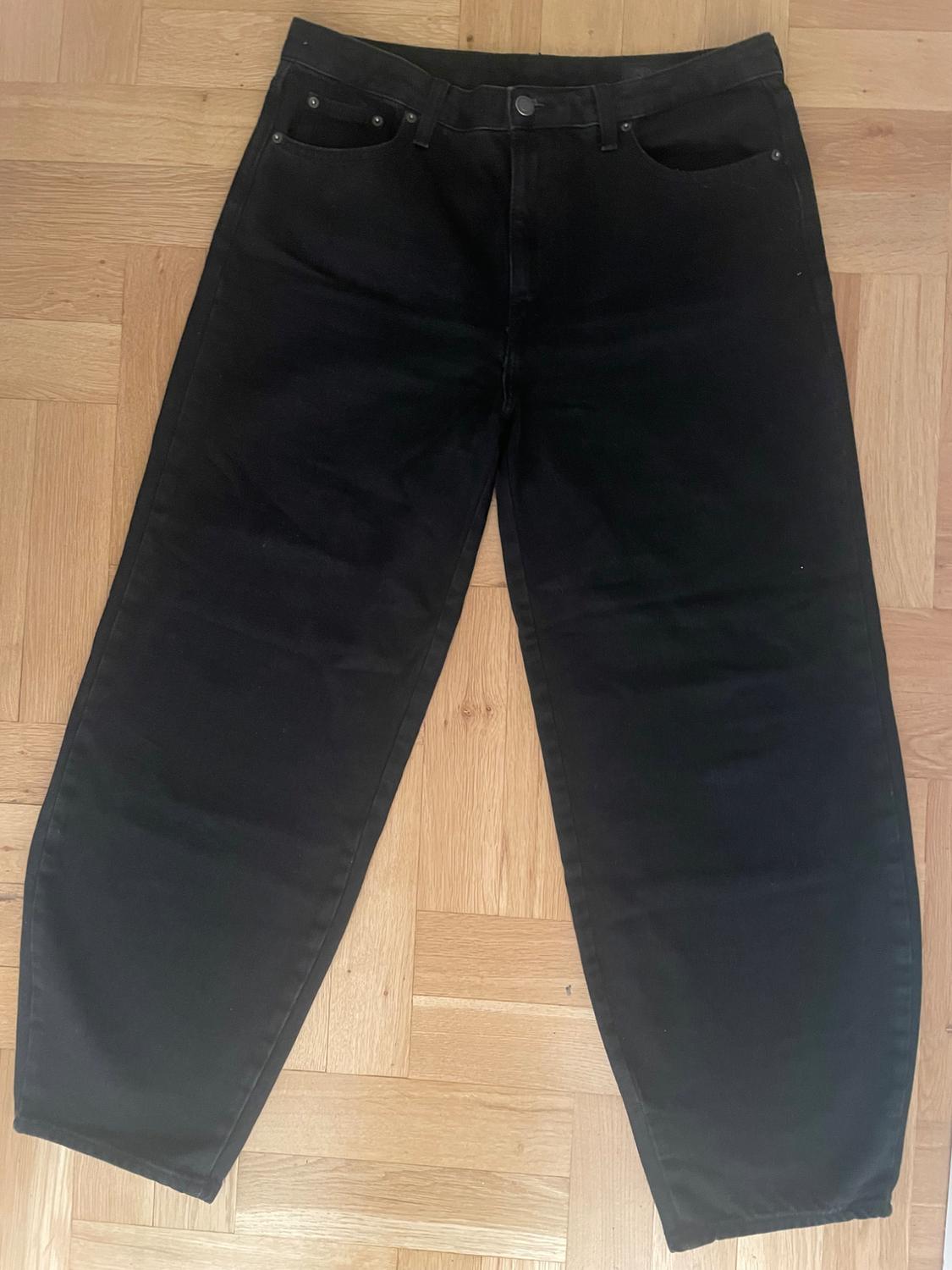 Black, straight-cut denim jeans, placed on a light wooden floor.