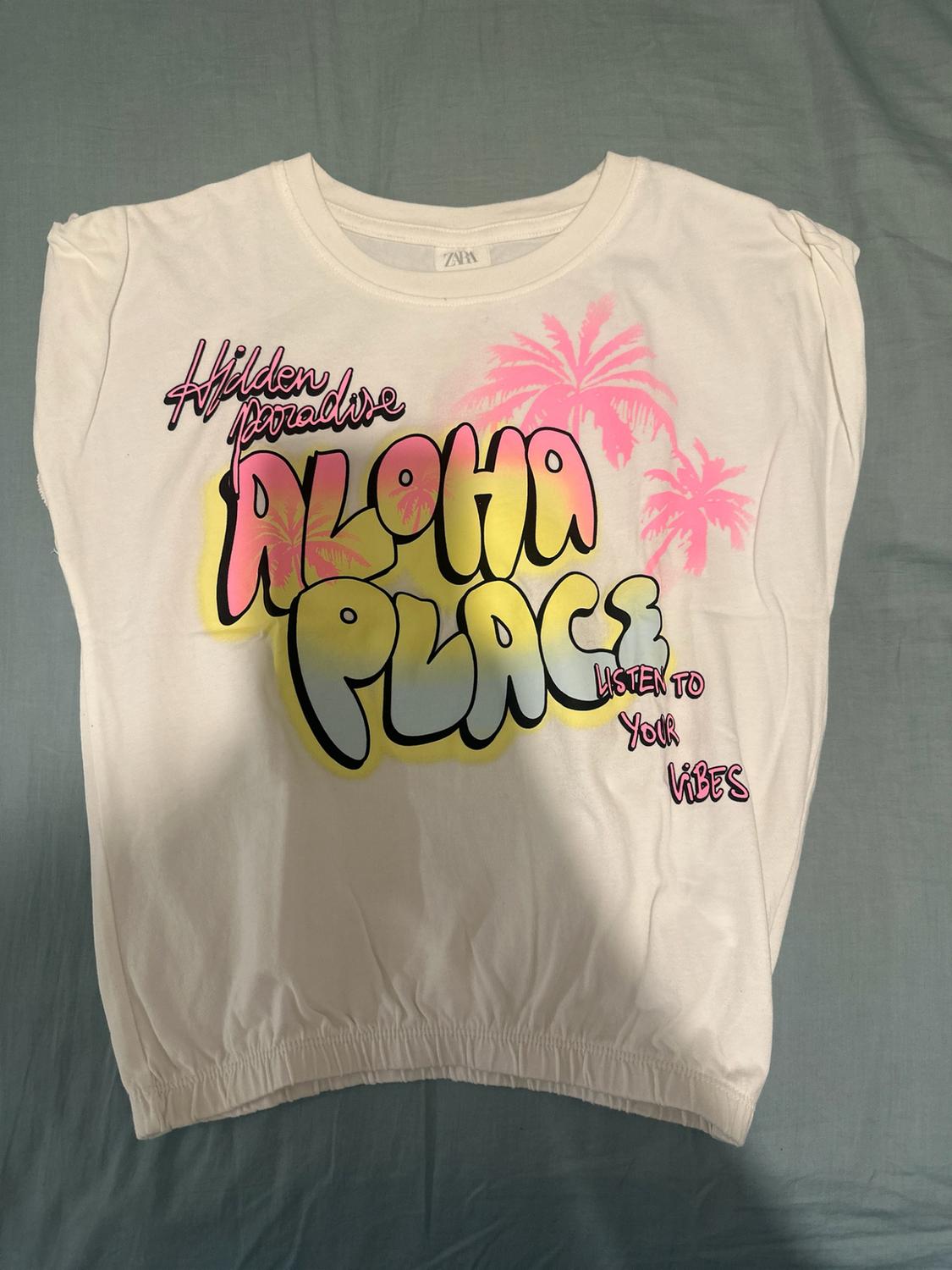 White sleeveless top with an elasticated hem and the text: Hidden Paradise ALOHA PLACE LISTEN TO YOUR VIBES, featuring pink palm trees.