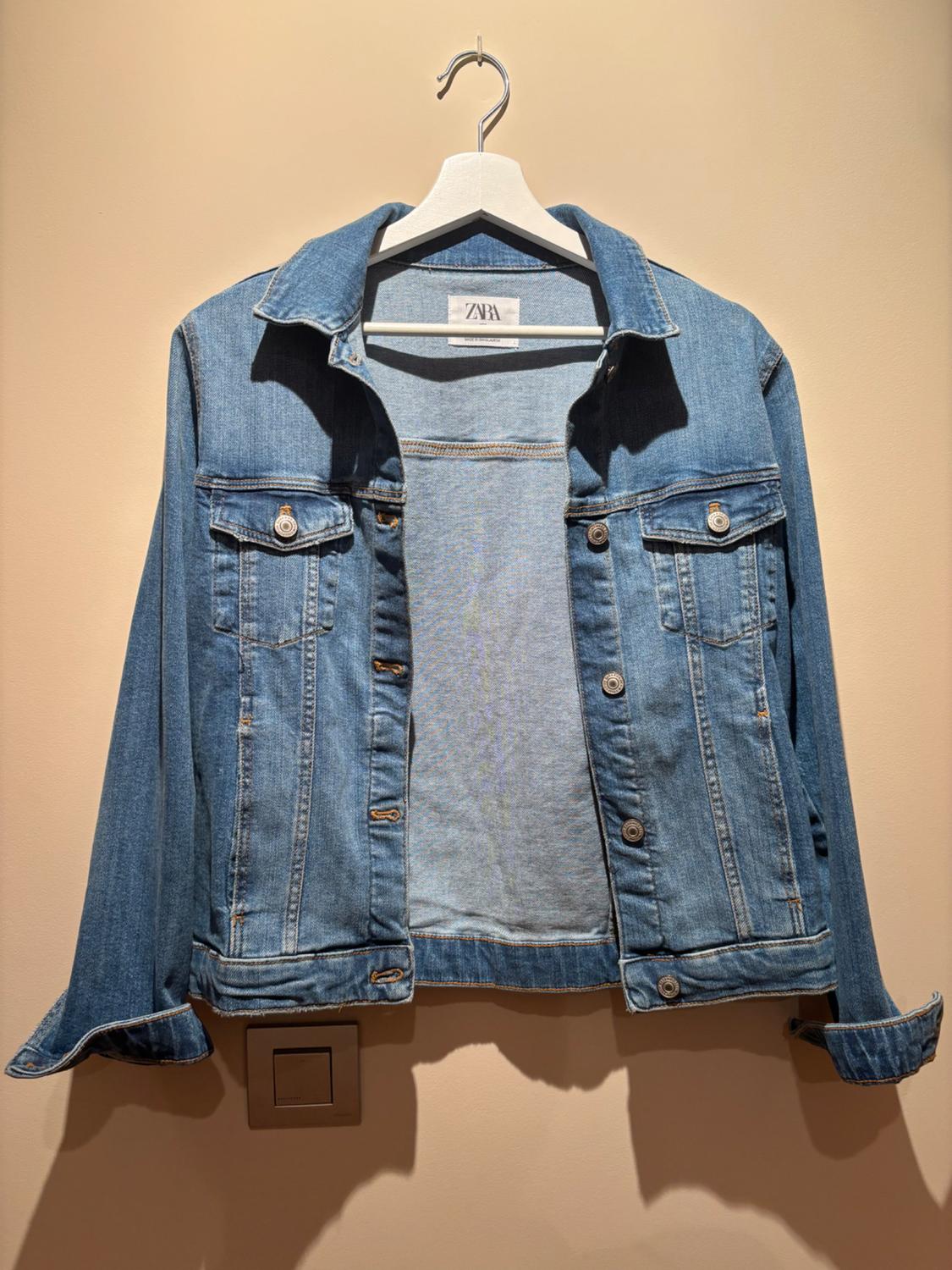 Blue denim jacket hanging on a white hanger against a beige wall, with the "ZARA" label visible inside the collar.