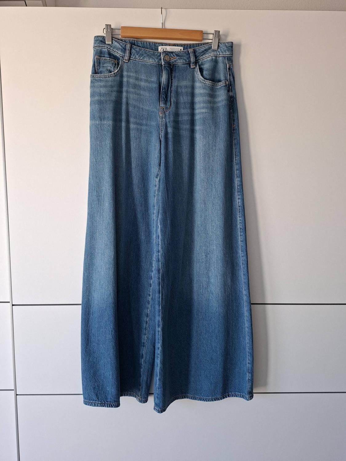 Blue wide-leg jeans with a distressed wash, hanging on a wooden hanger against a white background.
