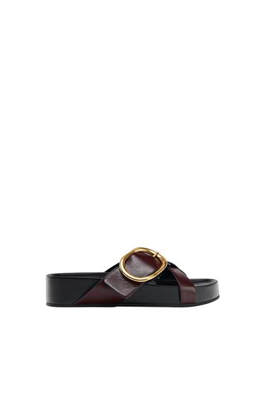 Black platform sandal with burgundy cross-over straps and a large circular gold buckle, side view on a white background.