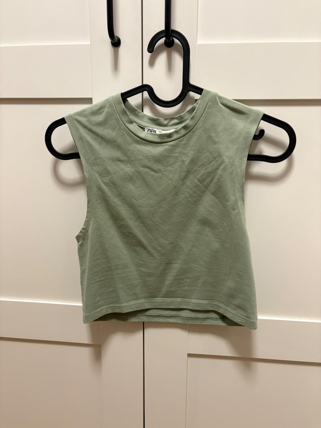Light green cropped sleeveless top hanging on a black hanger in front of a white wardrobe. The label reads ZARA.