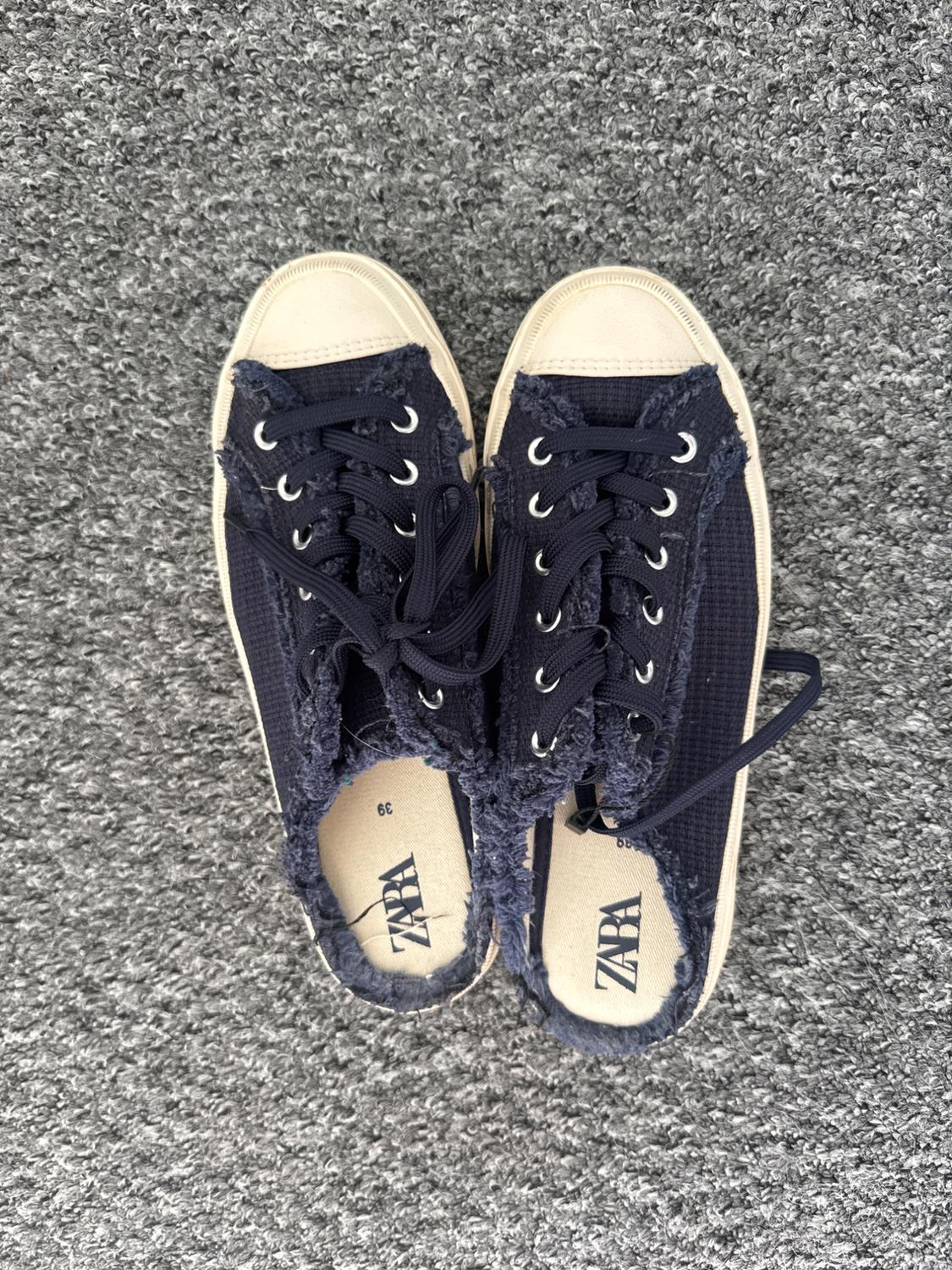 Navy canvas trainers with beige soles on a grey carpet. Visible text: "ZARA" and "39". Dark laces and frayed edge.
