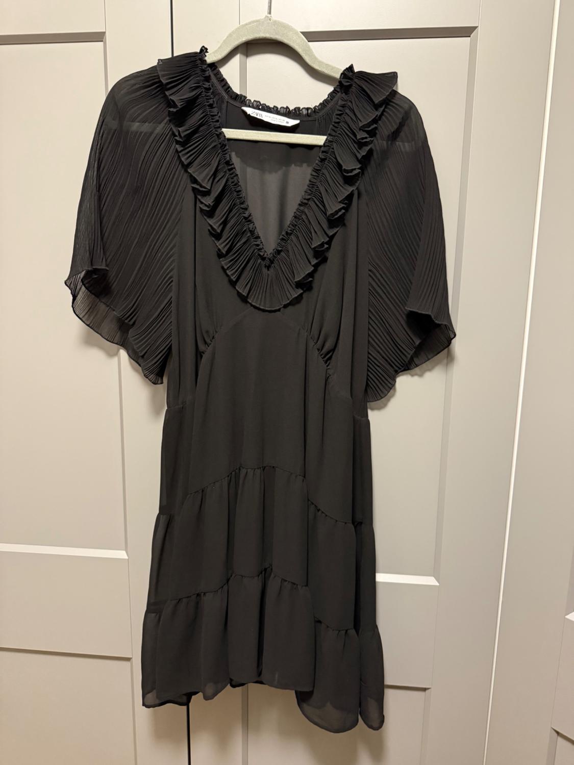 Short black dress with pleated sleeves and a ruffled V-neck, hanging on a white door. ZARA text on the tag.