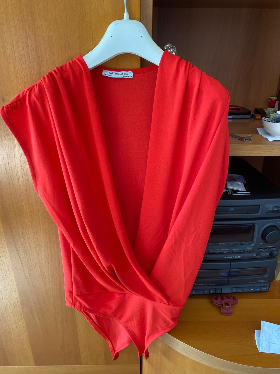 Red bodysuit with V-neckline hanging on a white hanger, against a wooden background and shelf with various objects.