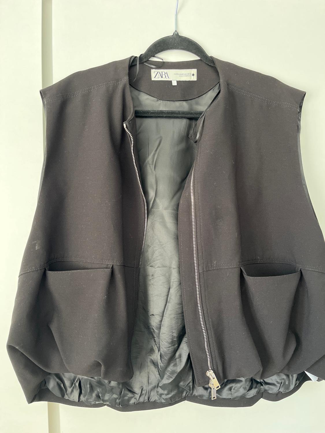 Black sleeveless gilet with a zip and large pockets, hanging on a black hanger. Visible label with the text "ZARA".