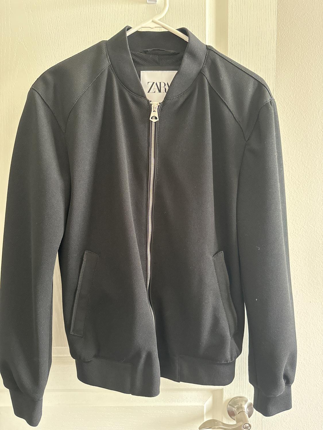 Black jacket with a metal zip, side pockets and a bomber-style collar, hanging on a hanger in front of a white door.