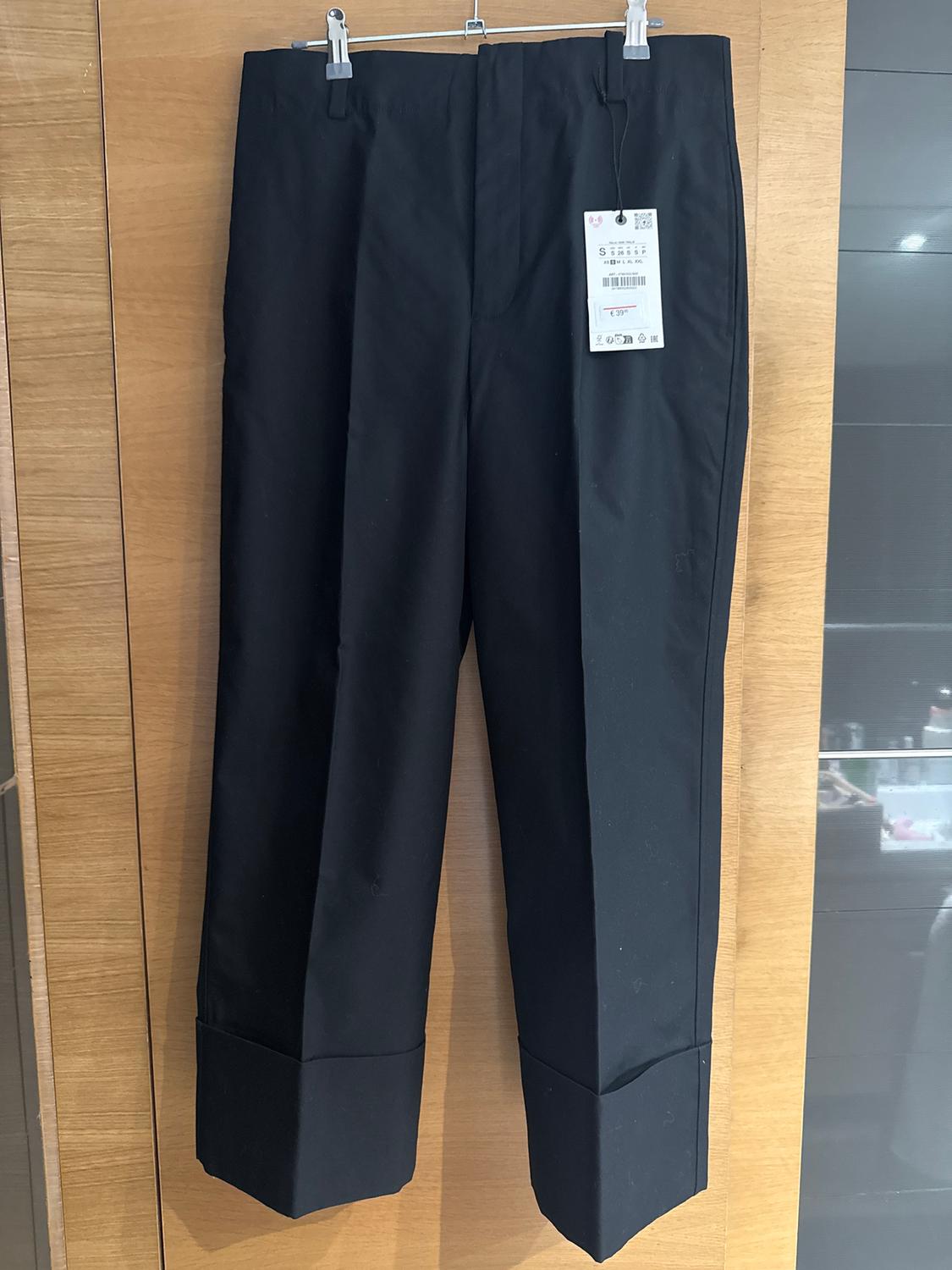 Black trousers hanging on a wooden hanger, with a visible tag. Light wood wall and dark panel background.