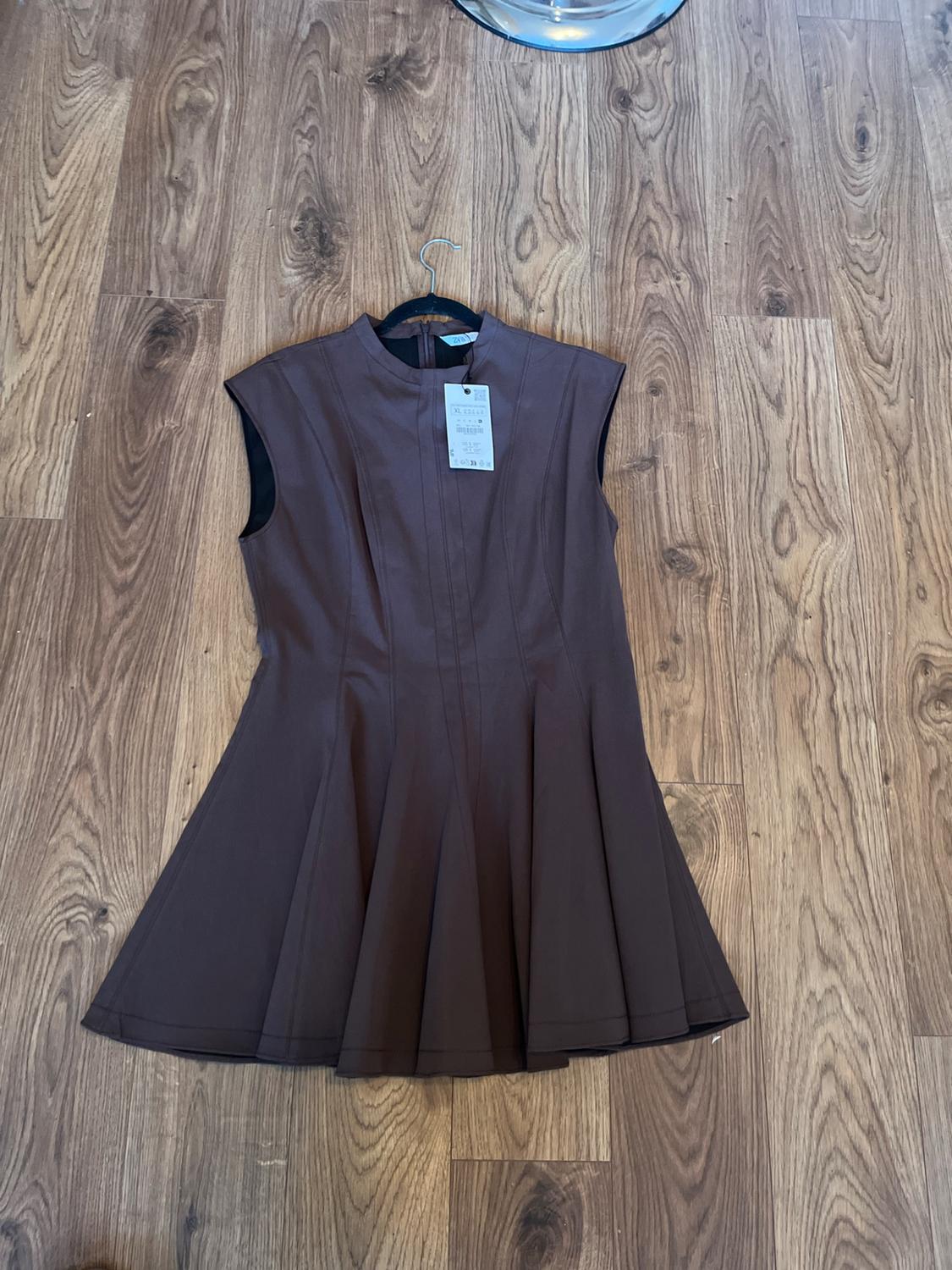 Brown sleeveless mini dress with a high neck and a flared skirt. It is hanging on a hanger with a tag against a wooden floor.