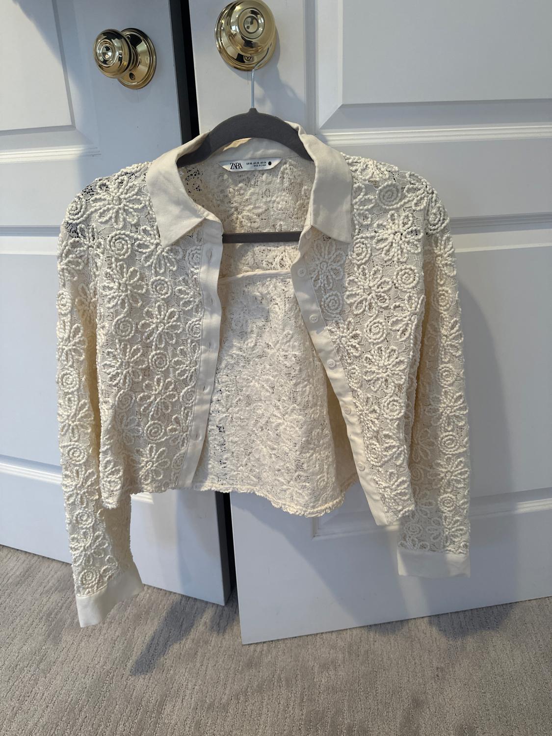 White lace jacket hanging on a hanger in front of a white door. The tag says "Zara".