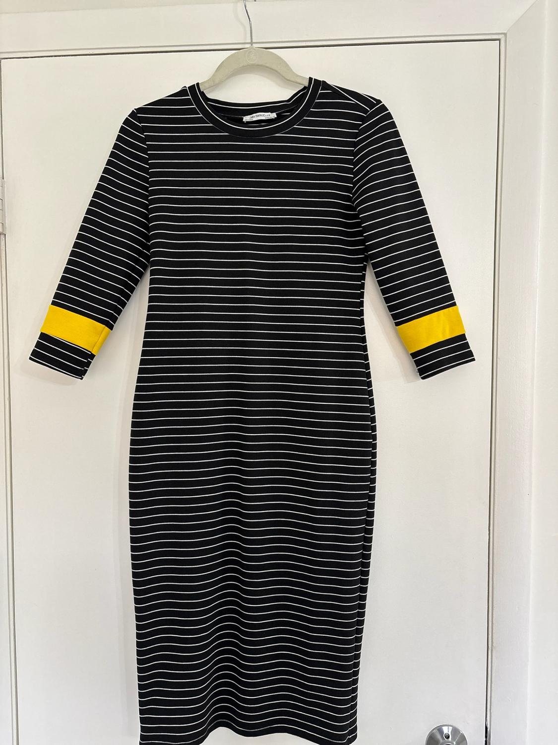Black dress with white horizontal stripes and a yellow band on the sleeves, hanging on a coat hanger over a white door.