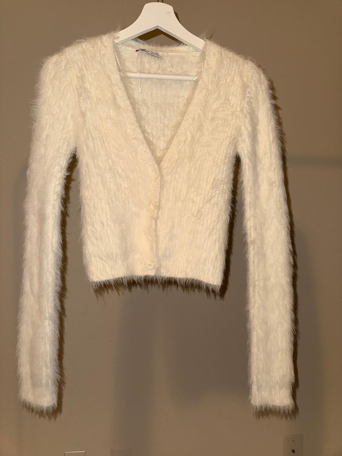 White fluffy textured cardigan hanging on a white hanger against a beige background.