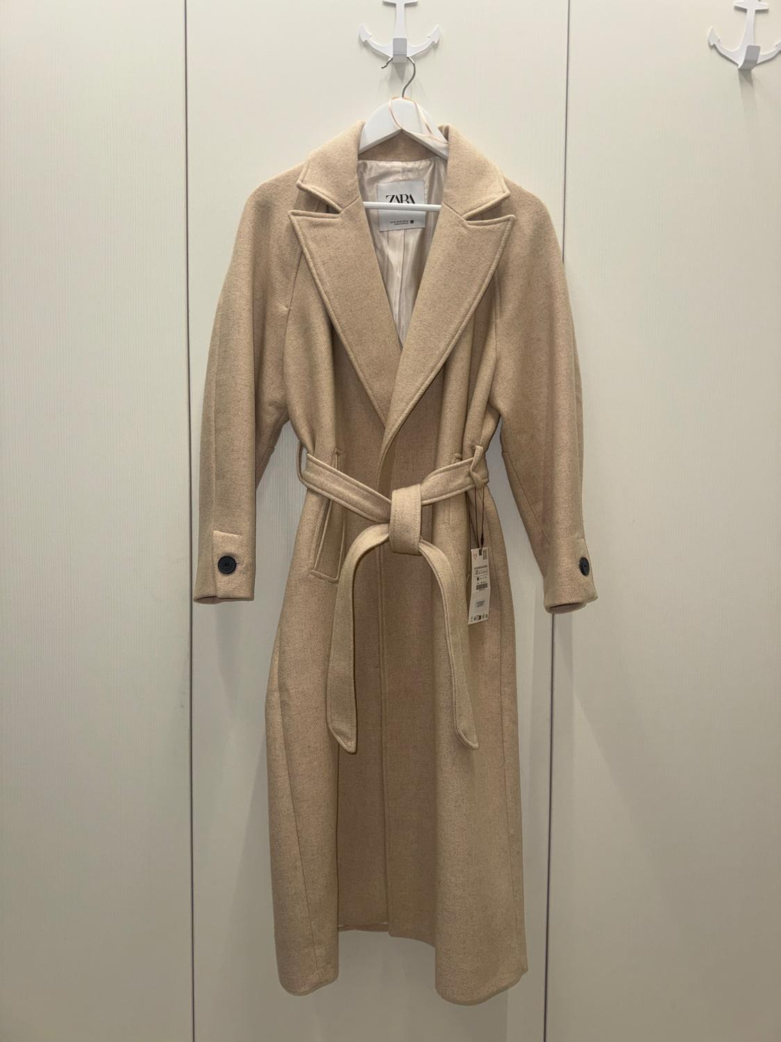 Beige coat with belt and wide lapels hanging on a white hanger. Visible label. White wall background with hooks.