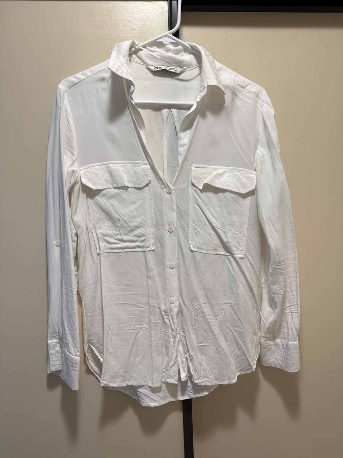 Long-sleeved white shirt with chest pockets, hanging on a hanger against a beige background. ZARA label visible.