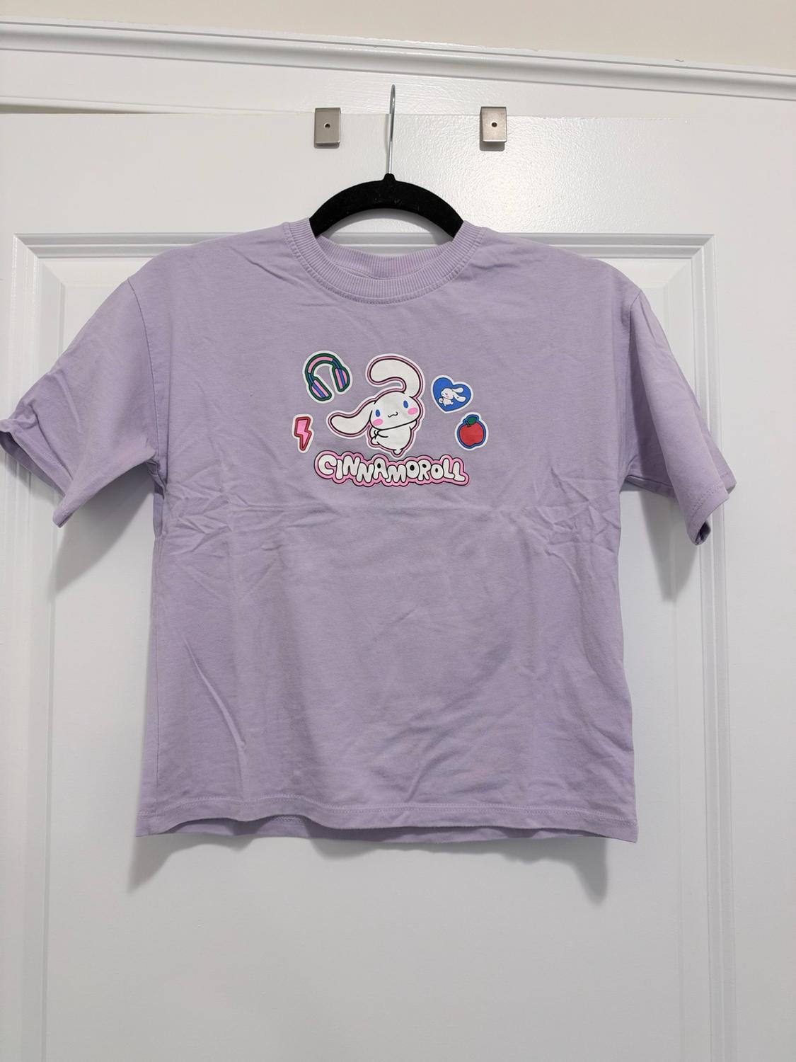 Lilac short-sleeved t-shirt hanging on a white door. It features a graphic print on the front with drawings and the text CINNAMOROLL.