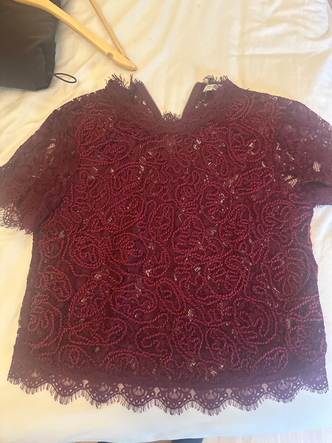 Burgundy short-sleeved lace top with a floral design, laid out on a white sheet next to a wooden hanger.
