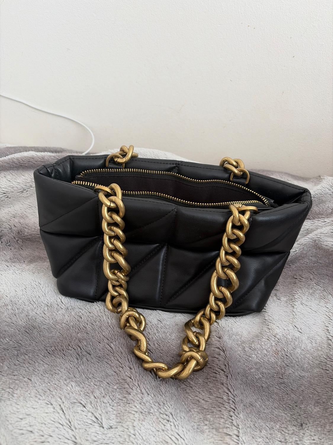 Black handbag with a quilted design and gold chain on a grey surface. White background.