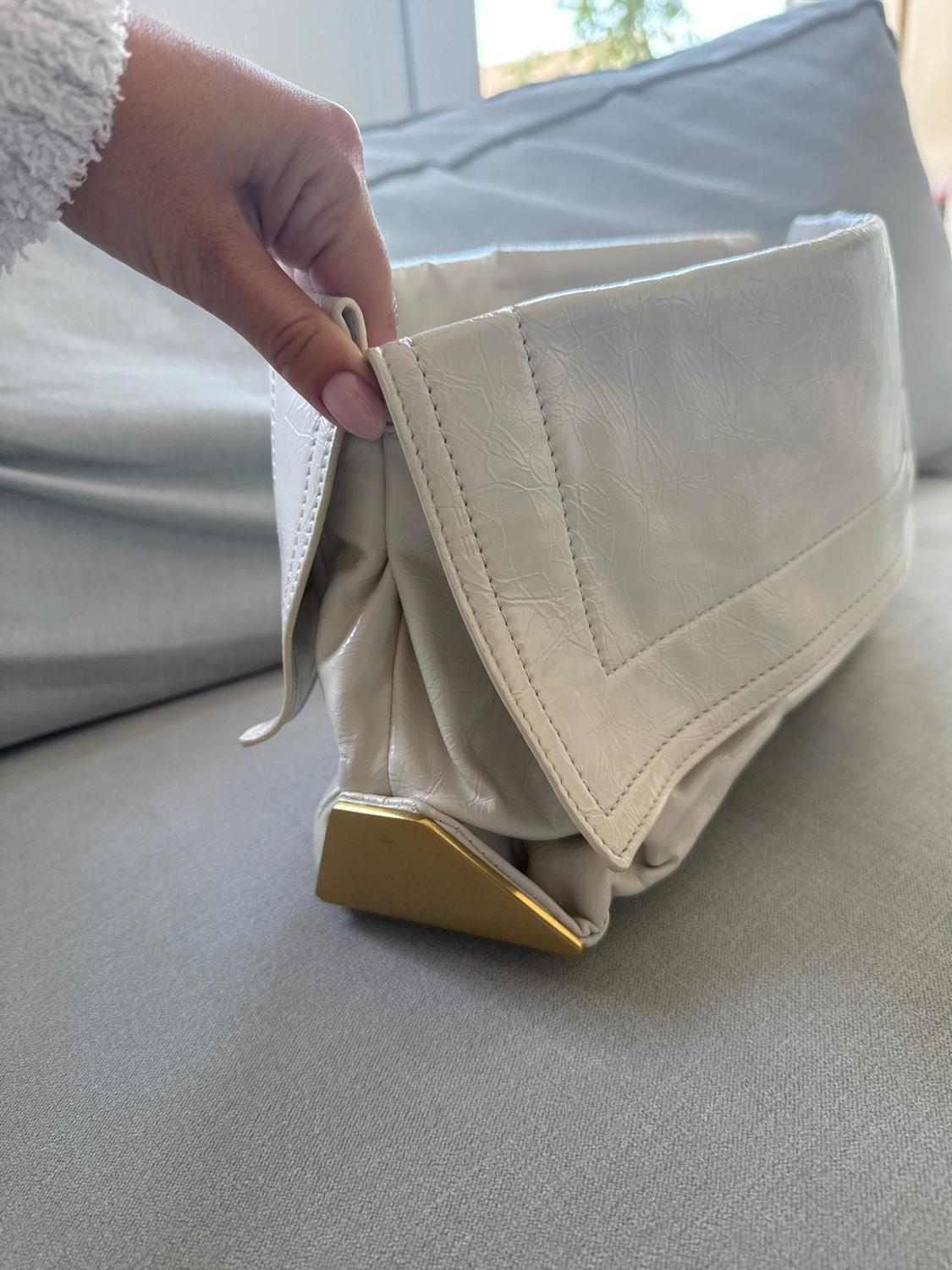 Hand holding a white crinkled-effect handbag with a flap and a gold metal corner, resting on a light grey sofa.