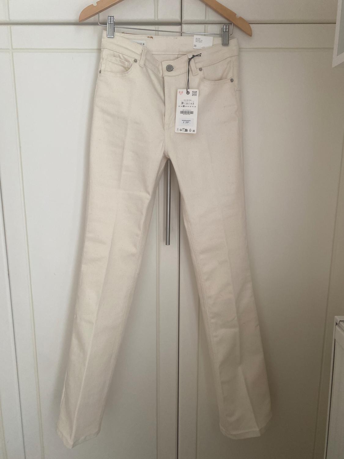White trousers hanging on a wooden hanger over a white door. Label visible on the waistband.