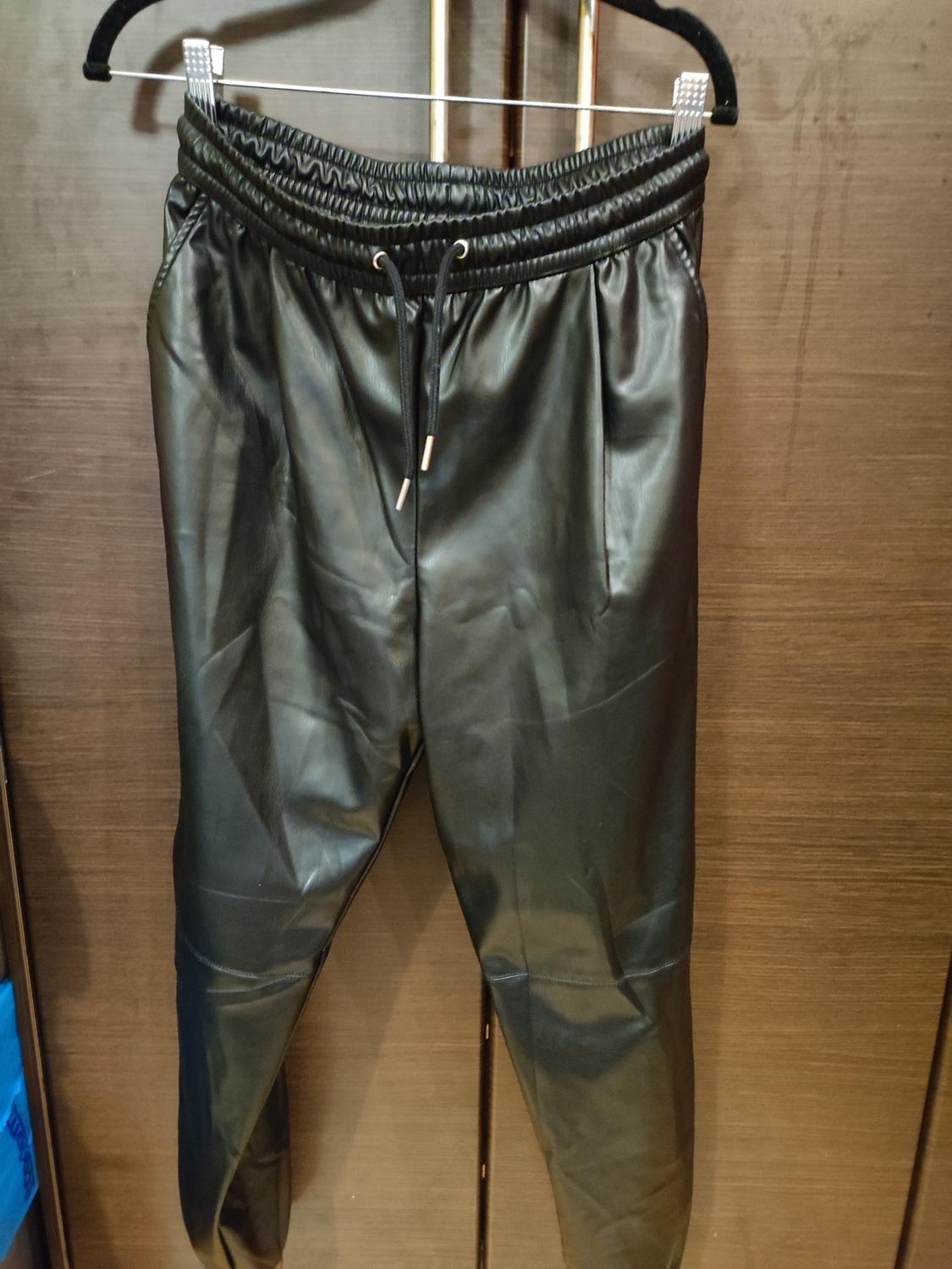 Black trousers made of shiny material hanging on a black hanger against a brown wooden background.