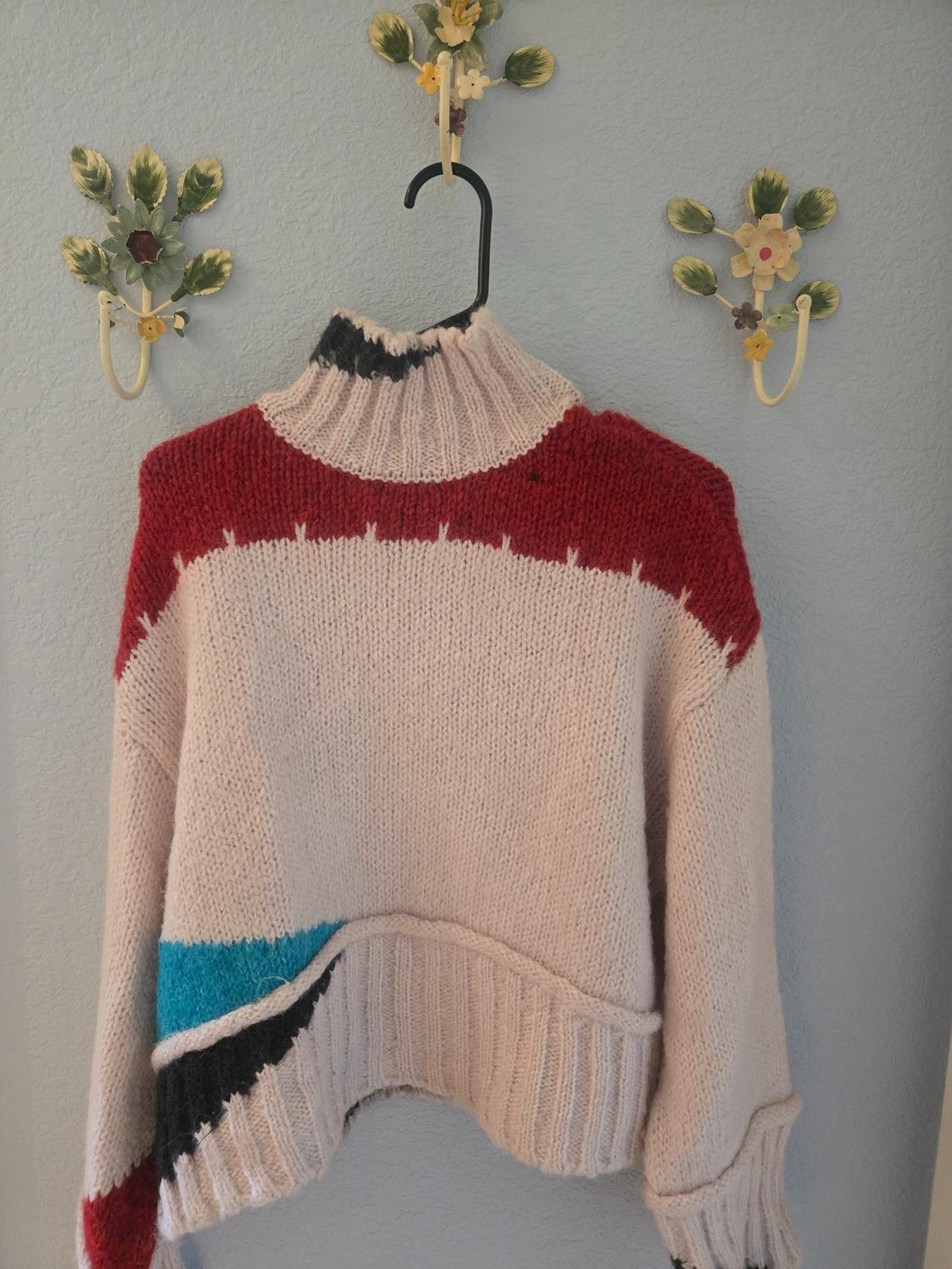 Knitted jumper hanging on a blue wall with decorative flower hooks. Red, black, and blue design on the collar and hem.
