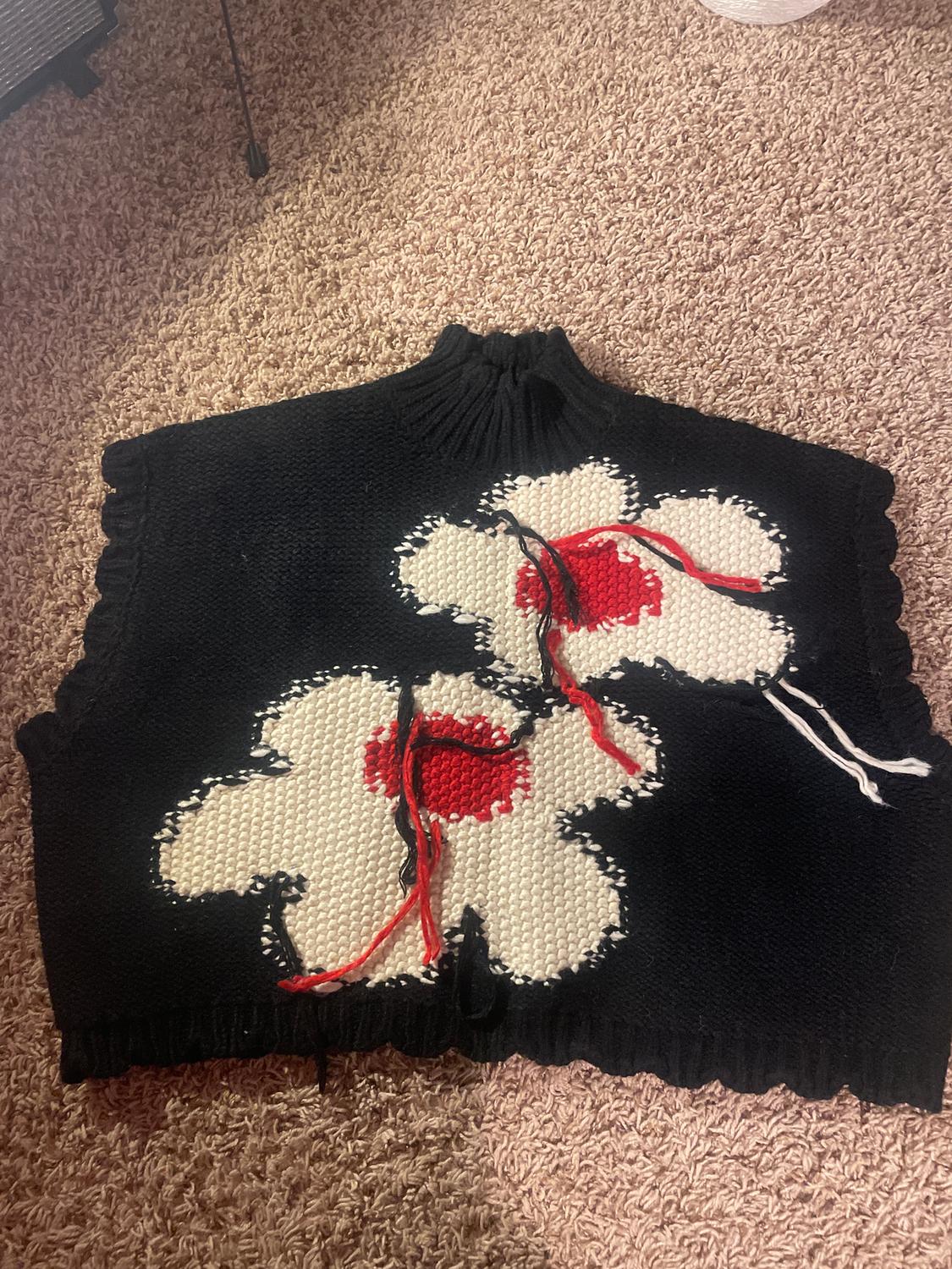 Black knitted waistcoat with a high neck, decorated with two red and white flowers. Beige carpet background.
