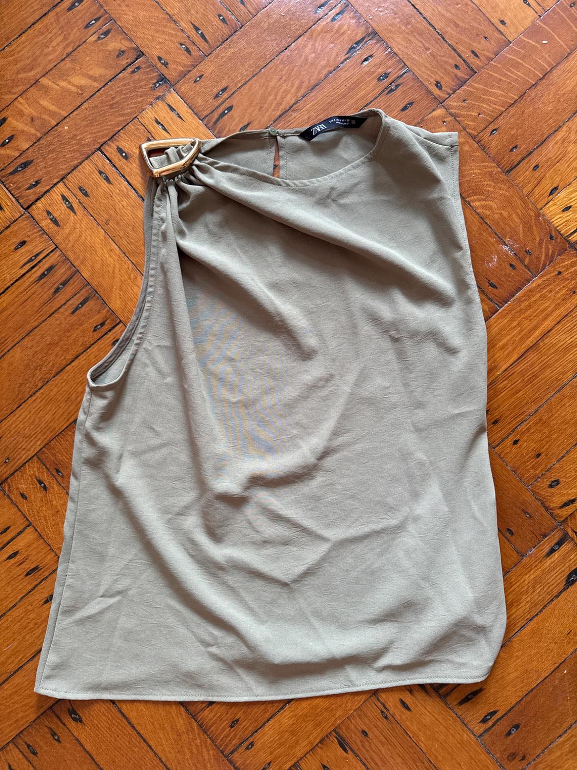 Khaki sleeveless blouse with a buckle detail on the shoulder, on a wooden floor with a geometric pattern.