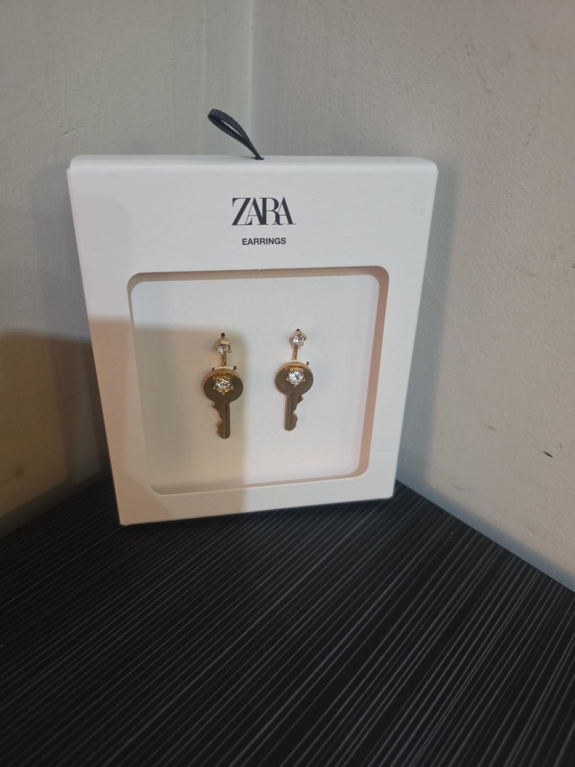 White box with the text "ZARA EARRINGS" containing gold-coloured key-shaped earrings with sparkling stones against a dark background.
