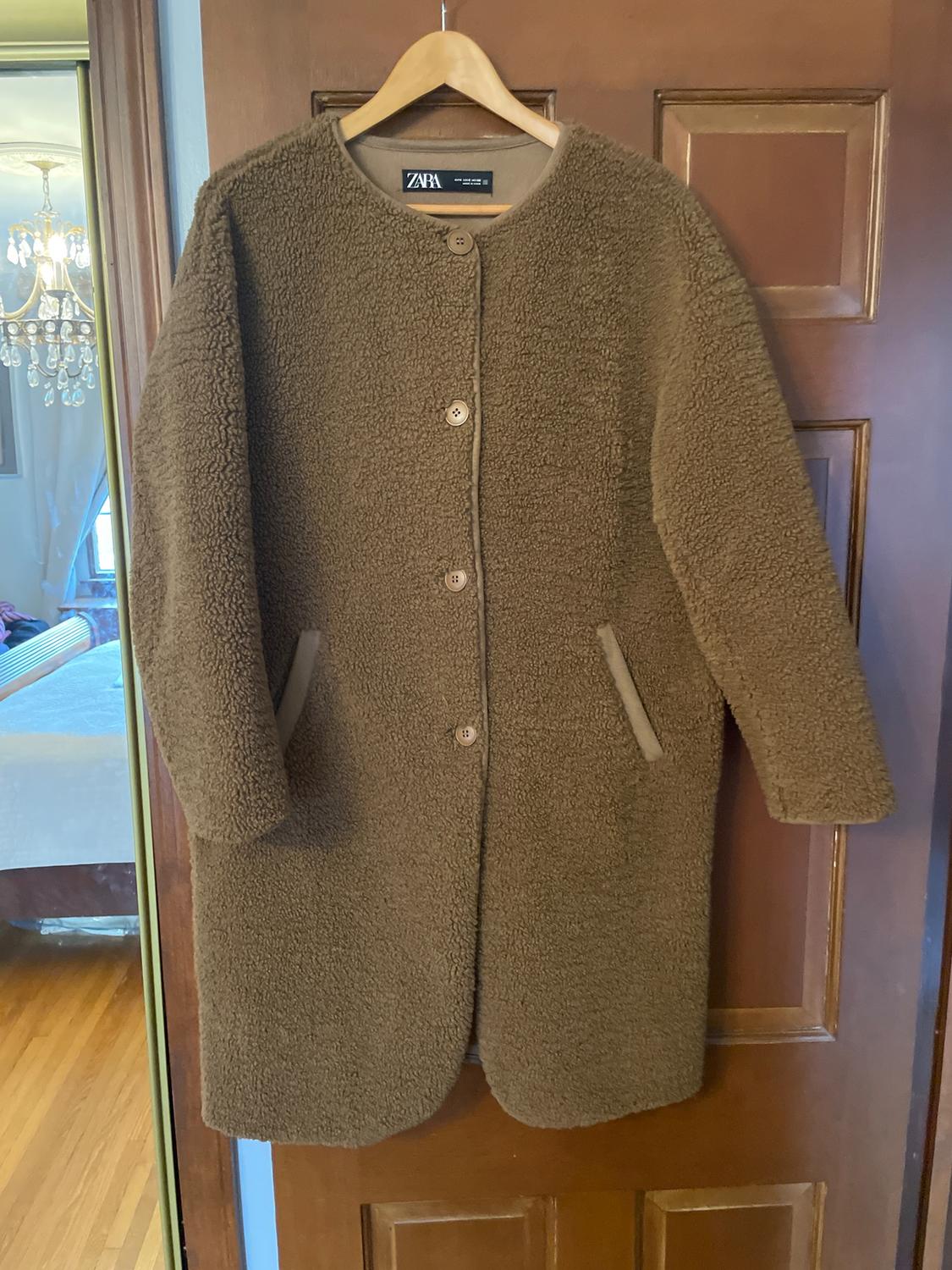Brown wool coat with buttons, hanging on a wooden door. Visible Zara tag. Background with lamp and room.