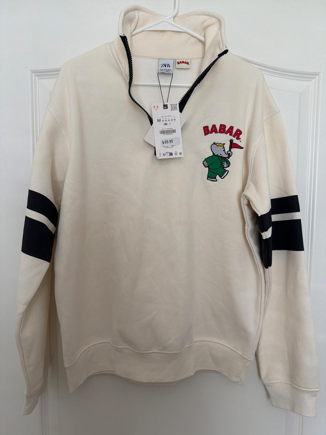 White zippered sweatshirt with green elephant drawing. Red "BABAR" text. Price tag $69.90 hanging.