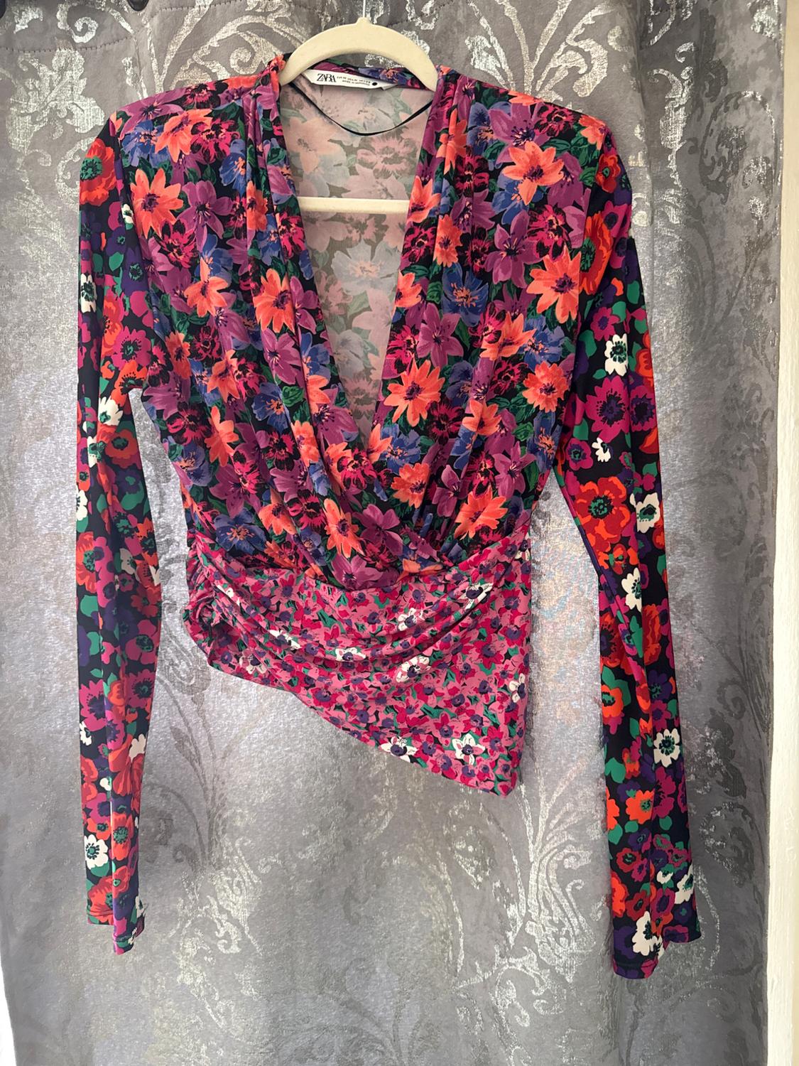 Long-sleeved blouse with a floral print in shades of pink, purple, and green, hanging on a hanger against a gray patterned background.