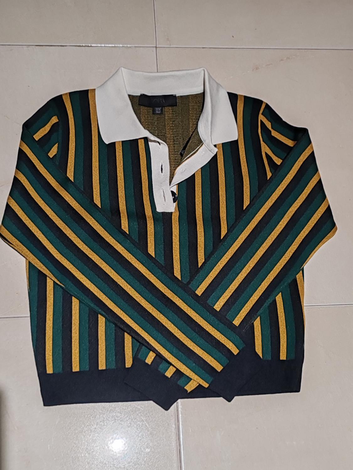 Green and yellow striped jumper with a white collar on a light-coloured floor. Sleeves crossed at the front.
