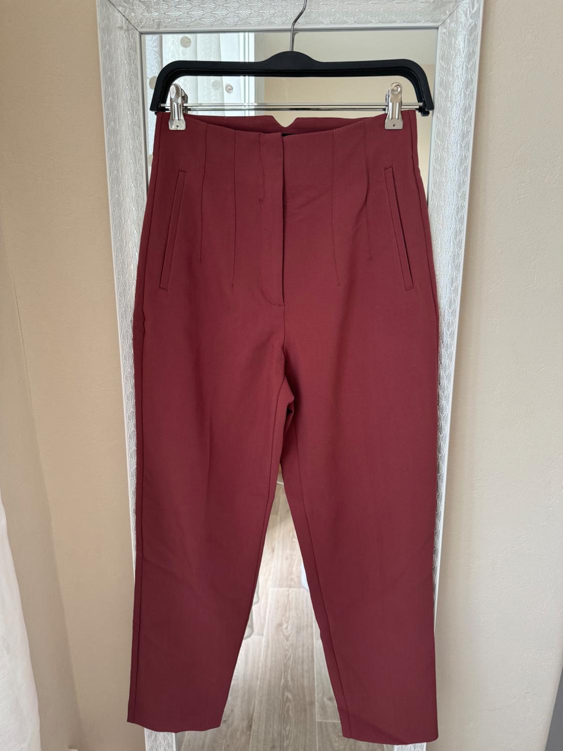 Burgundy trousers hanging on a black hanger over a white-framed mirror, in a well-lit indoor setting.